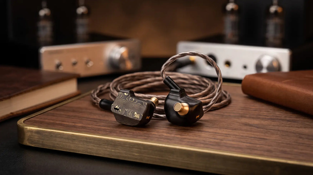 Campfire Audio audiophile in-ear monitors with black damascus texture and gold brass accents on a wooden desk.